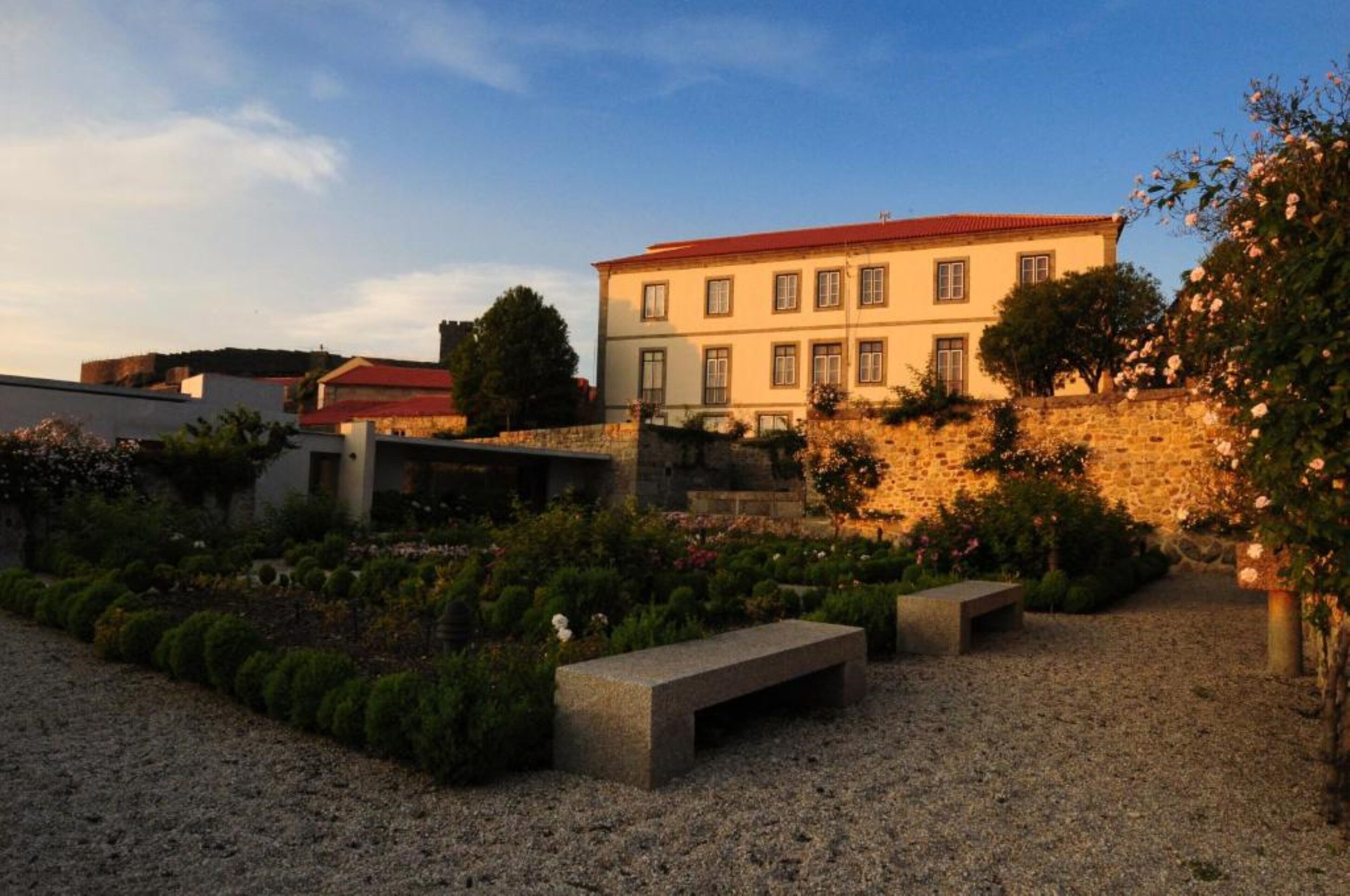 A courtyard with a stone wall, a bench, and a building with a red roof.