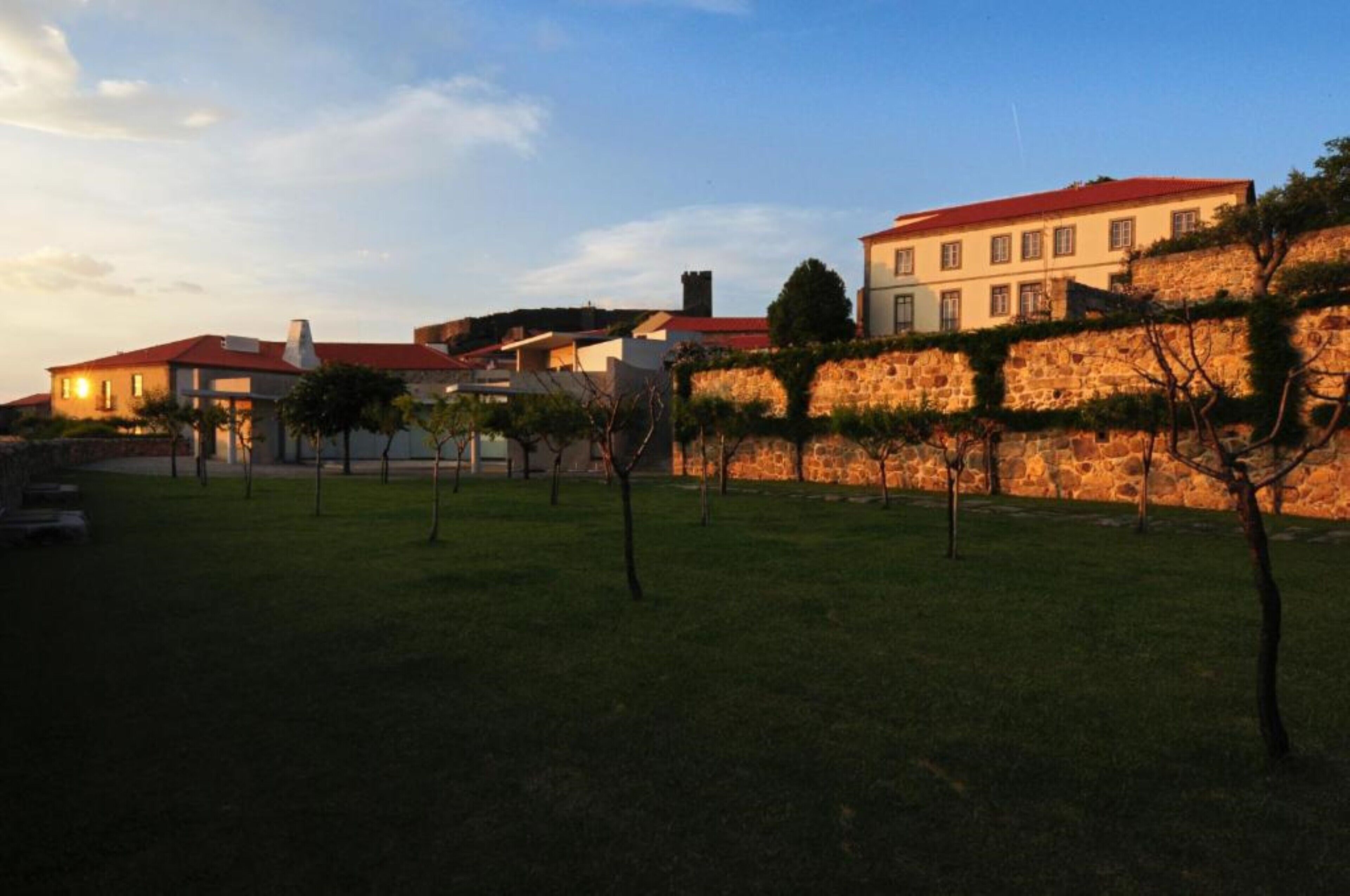 Un edificio grande con techo rojo, rodeado por un muro de piedra y una zona verde con árboles.