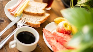 Une table en bois dressée pour le petit-déjeuner, avec du pain, du jambon, du fromage, une pomme, une pastèque et une tasse de café.