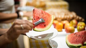 Une tranche de pastèque sur une assiette blanche, accompagnée d’une louche en métal, le tout entouré de pommes et d’autres fruits sur une table.