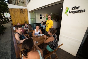 An outdoor dining area with a group of people seated at a wooden table, a man standing in the doorway, and a sign with a lizard logo.