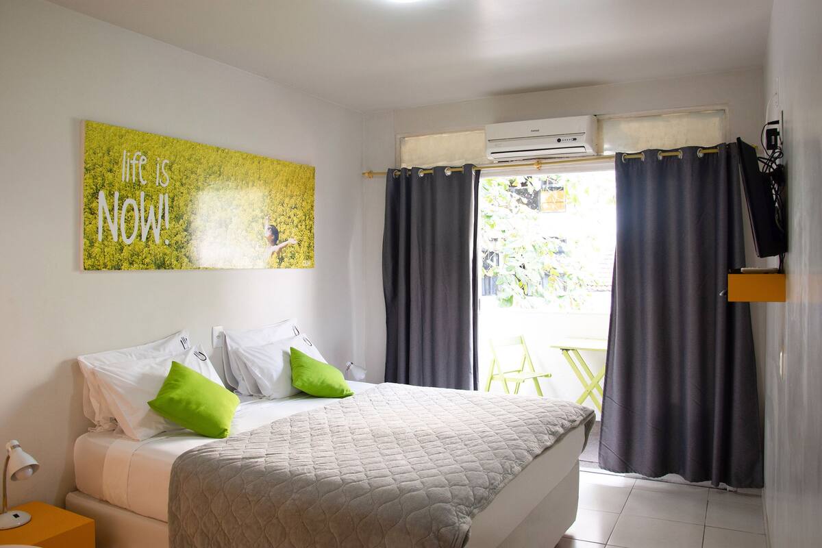 Une chambre à coucher avec un lit, un oreiller vert, un oreiller blanc, une couvre-lit matelassée grise, un climatiseur fixé au mur et une fenêtre avec des rideaux foncés.