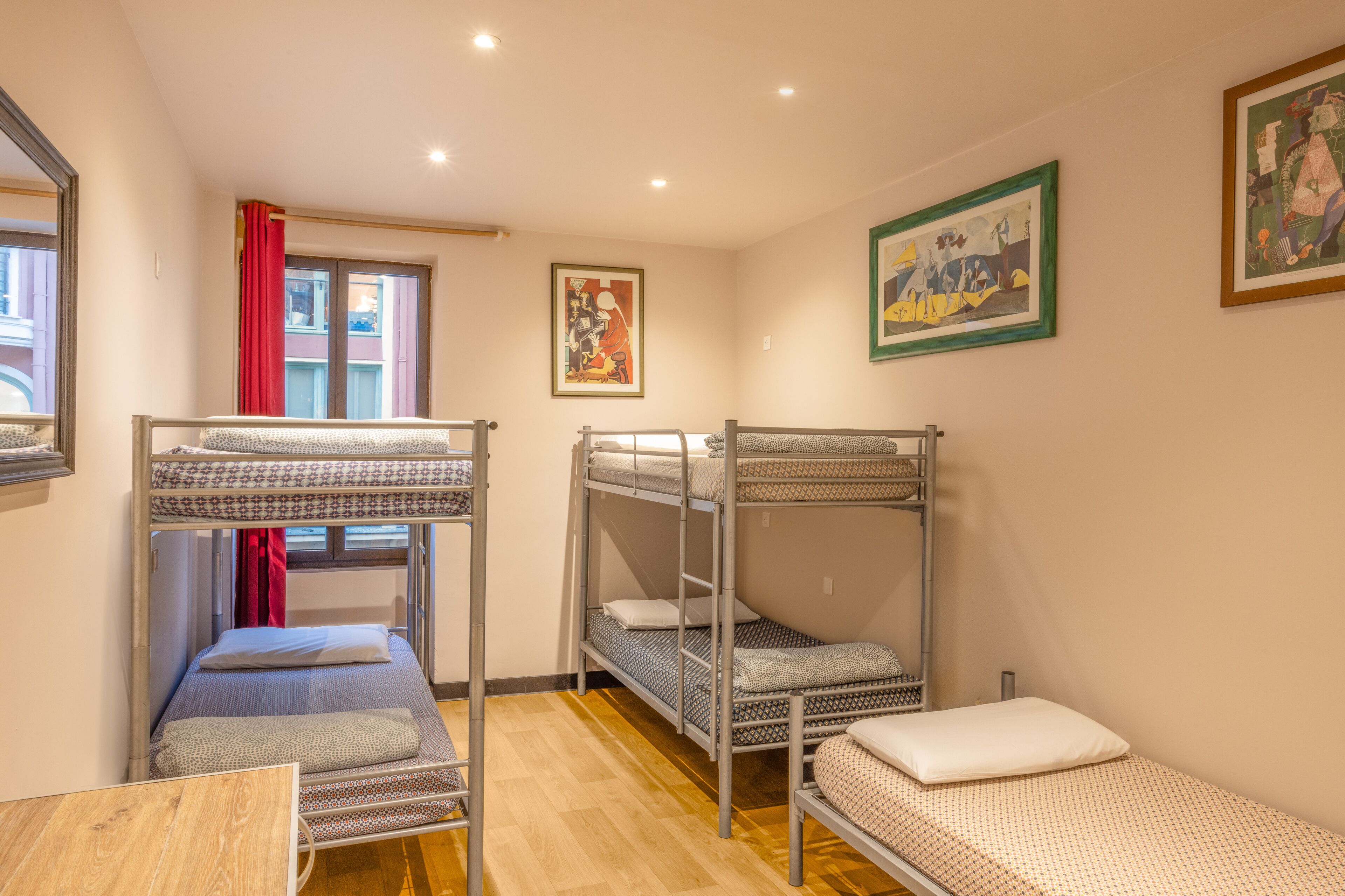 A dormitory room with bunk beds, a window with red curtains, and two paintings on the walls.