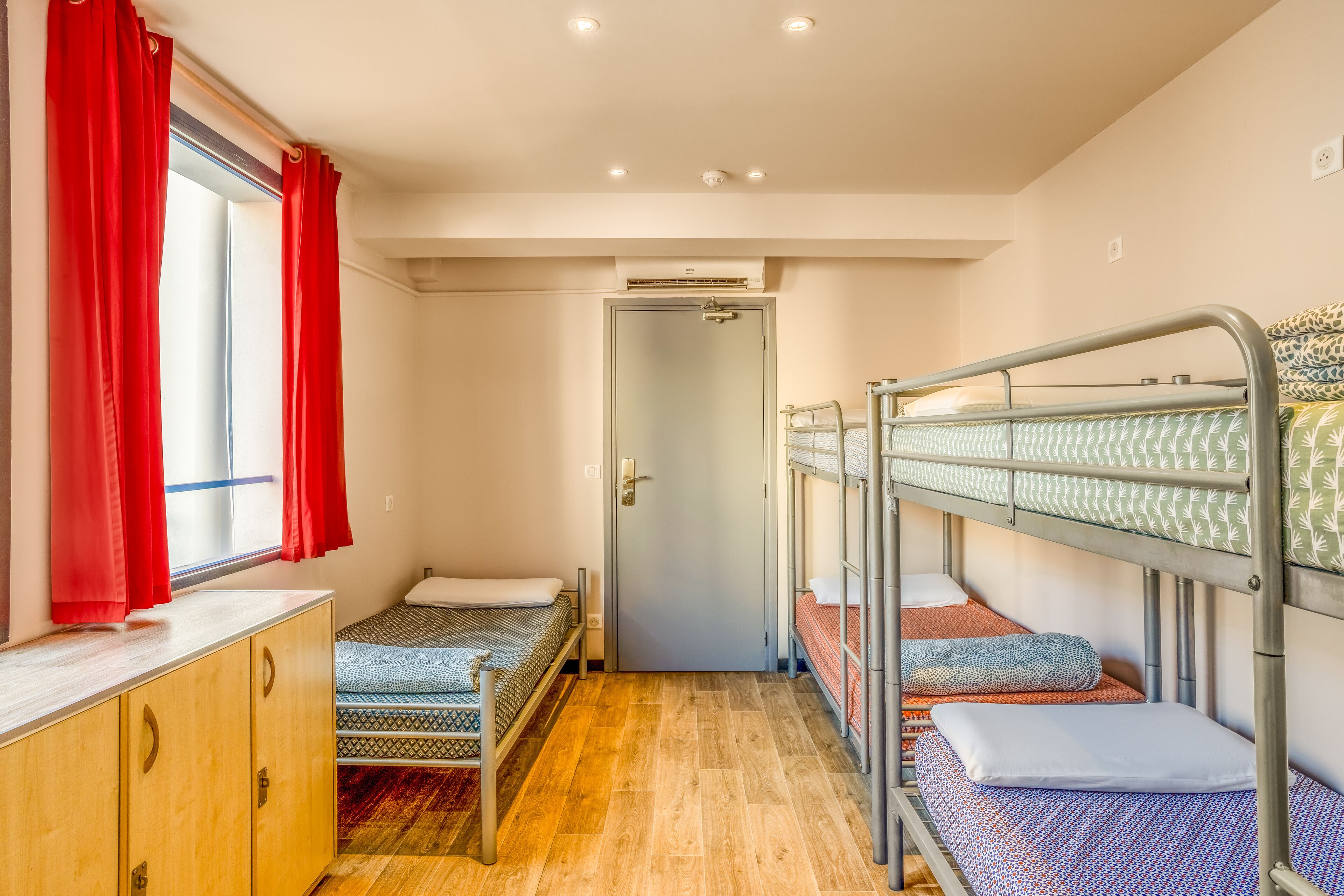 A dormitory room with bunk beds, a wooden wardrobe, a window with red curtains, and a door with an air conditioning unit above it.