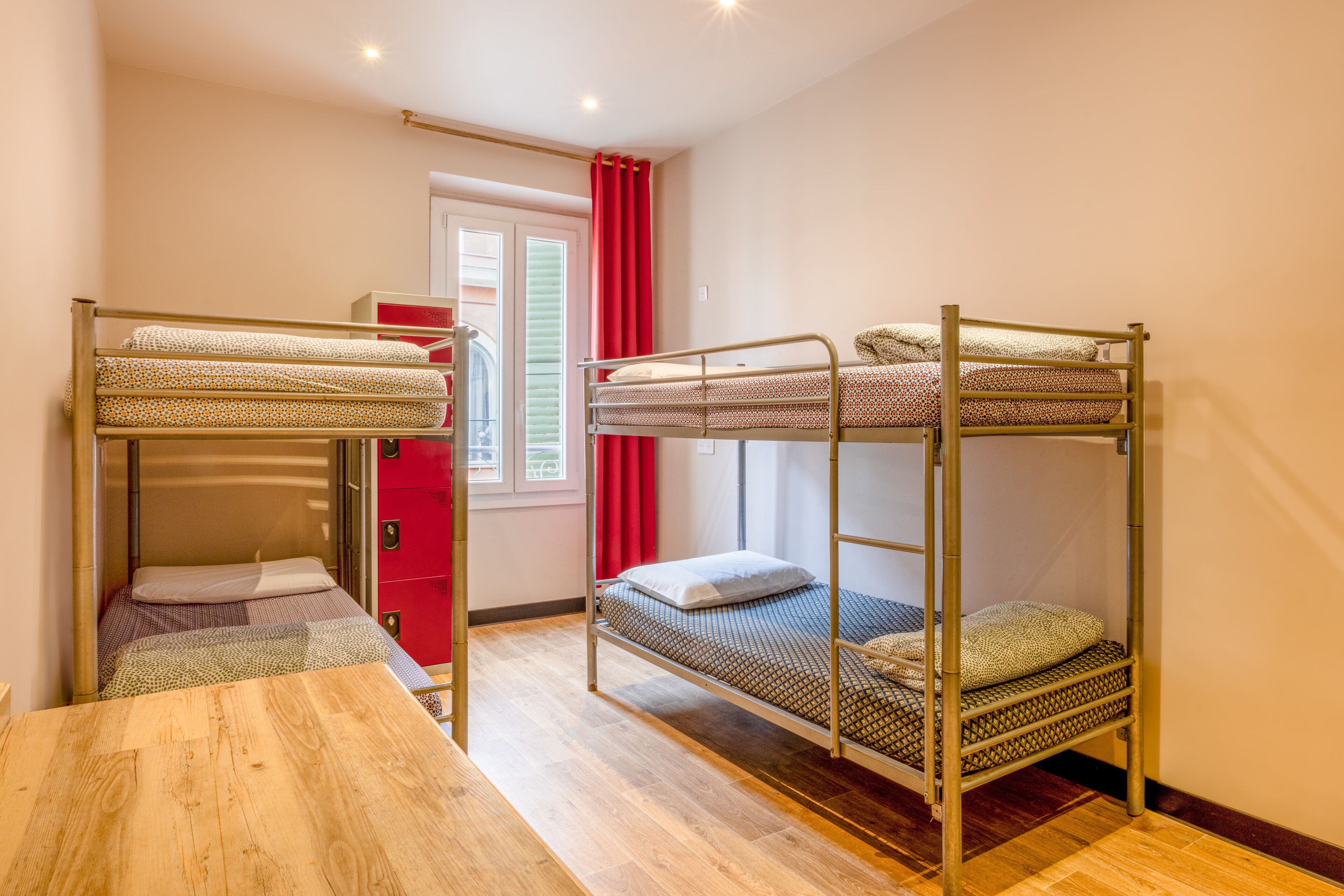 Una habitación tipo dormitorio con tres literas, una mesa de madera, un armario rojo y una ventana con cortinas rojas.