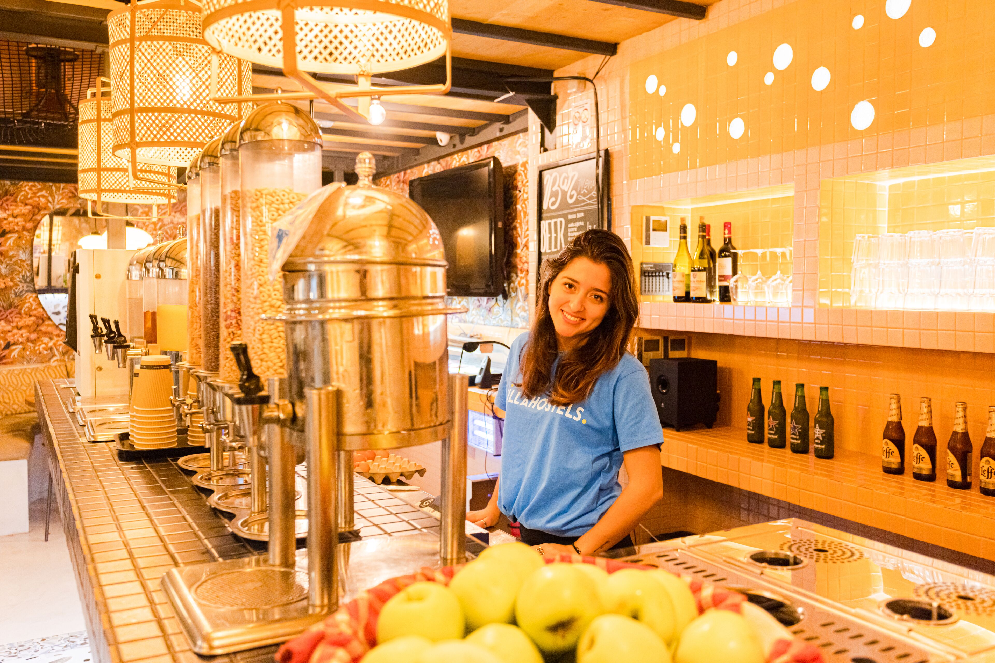 A bar area with a woman standing behind the counter, a display of bottles, and a basket of apples.