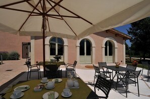 Un espace de restauration en plein air, avec des tables et des chaises disposées sous un grand parasol, le tout adossé à un bâtiment doté d’arcades et d’une façade rose.