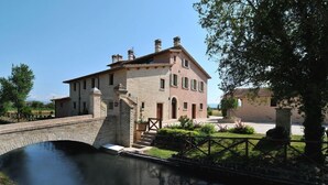 Un bâtiment en pierre de deux étages, surmonté d’un pont qui enjambe un canal, entouré de verdure et sous un ciel dégagé.