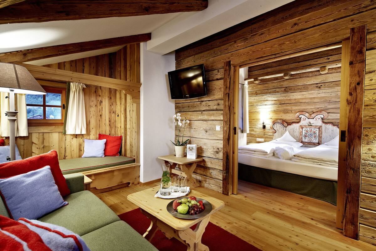 A cozy cabin-style room with a wooden interior, a bed with a decorative headboard, a television mounted on the wall, and a seating area with cushions and a table with fruit.