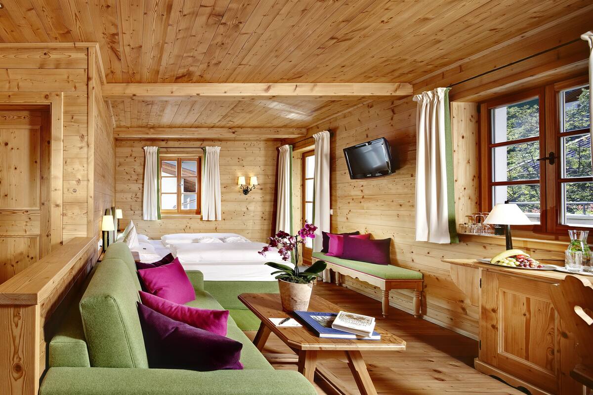 A wooden cabin interior with a bed, green and purple sofa, a dining table with a book and a plant, and a TV mounted on the wall.