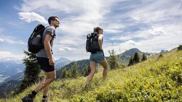 Two hikers on a grassy mountain trail with a scenic view of distant mountains and a partly cloudy sky.