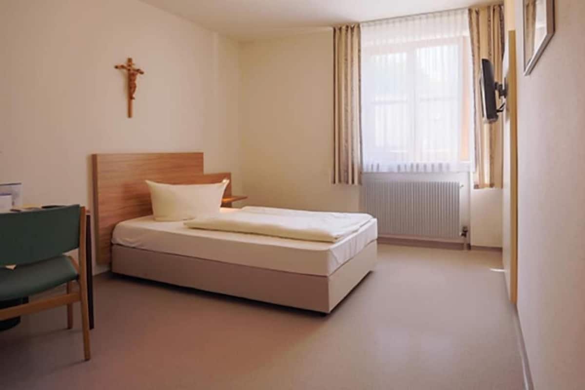 A hotel room with a bed, a chair, a cross on the wall, and a window with curtains.