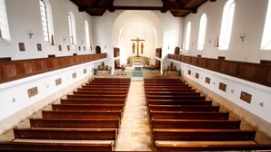 A church interior with wooden pews arranged in rows, a central altar, and arched windows.