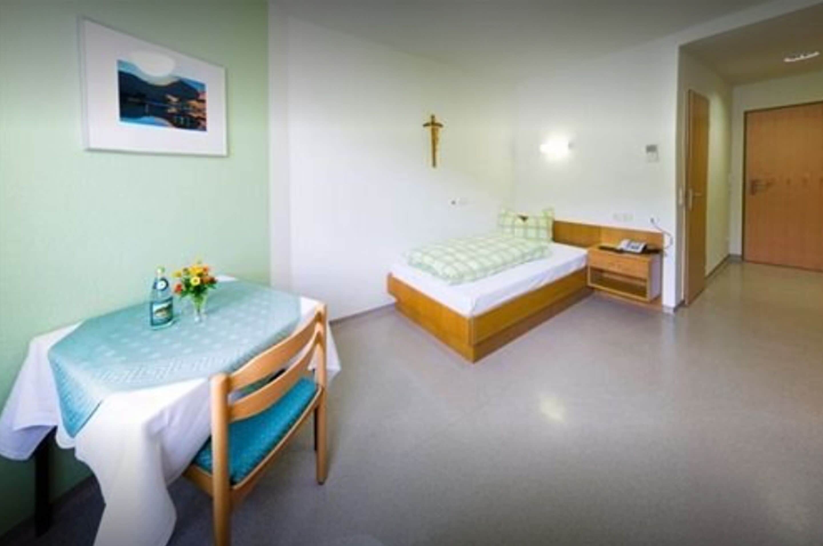 Ein kleiner, sauberer Raum mit einem Bett, einem Nachttisch und einem Stuhl. An der Wand hängt ein gerahmtes Bild und ein Kruzifix.