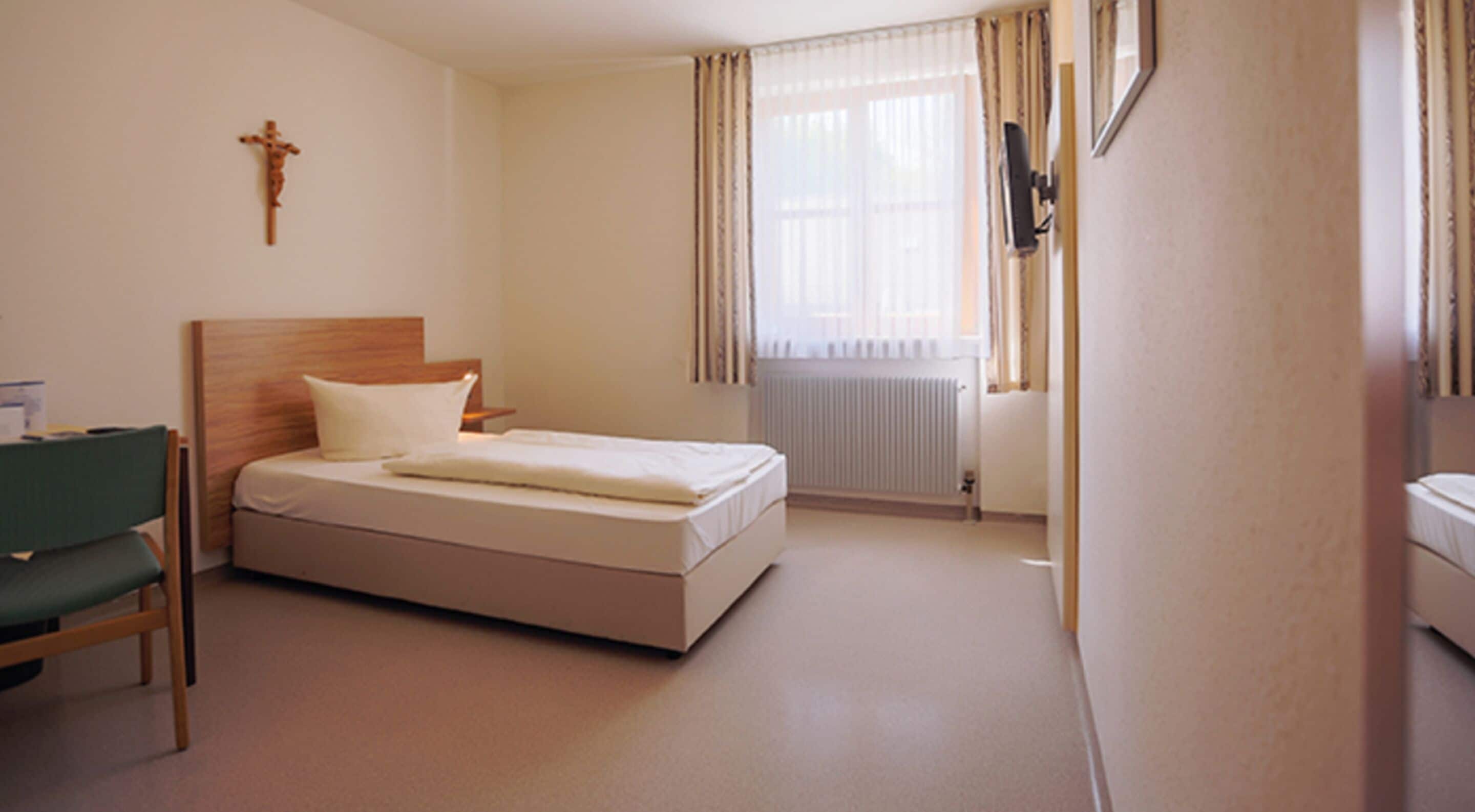 A hotel room with a single bed, a wooden headboard, a green chair, a bedside table, a wall-mounted TV, and a radiator.