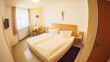 A hotel room with a large bed, a desk with a chair, a small table, a cross on the wall, and a door.