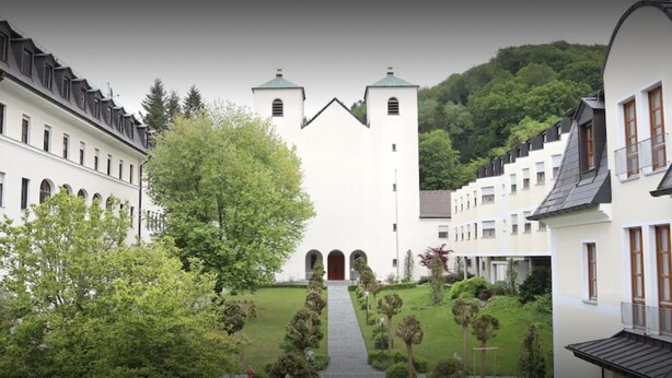A courtyard with a central building featuring two towers, surrounded by trees and a pathway leading to the entrance.