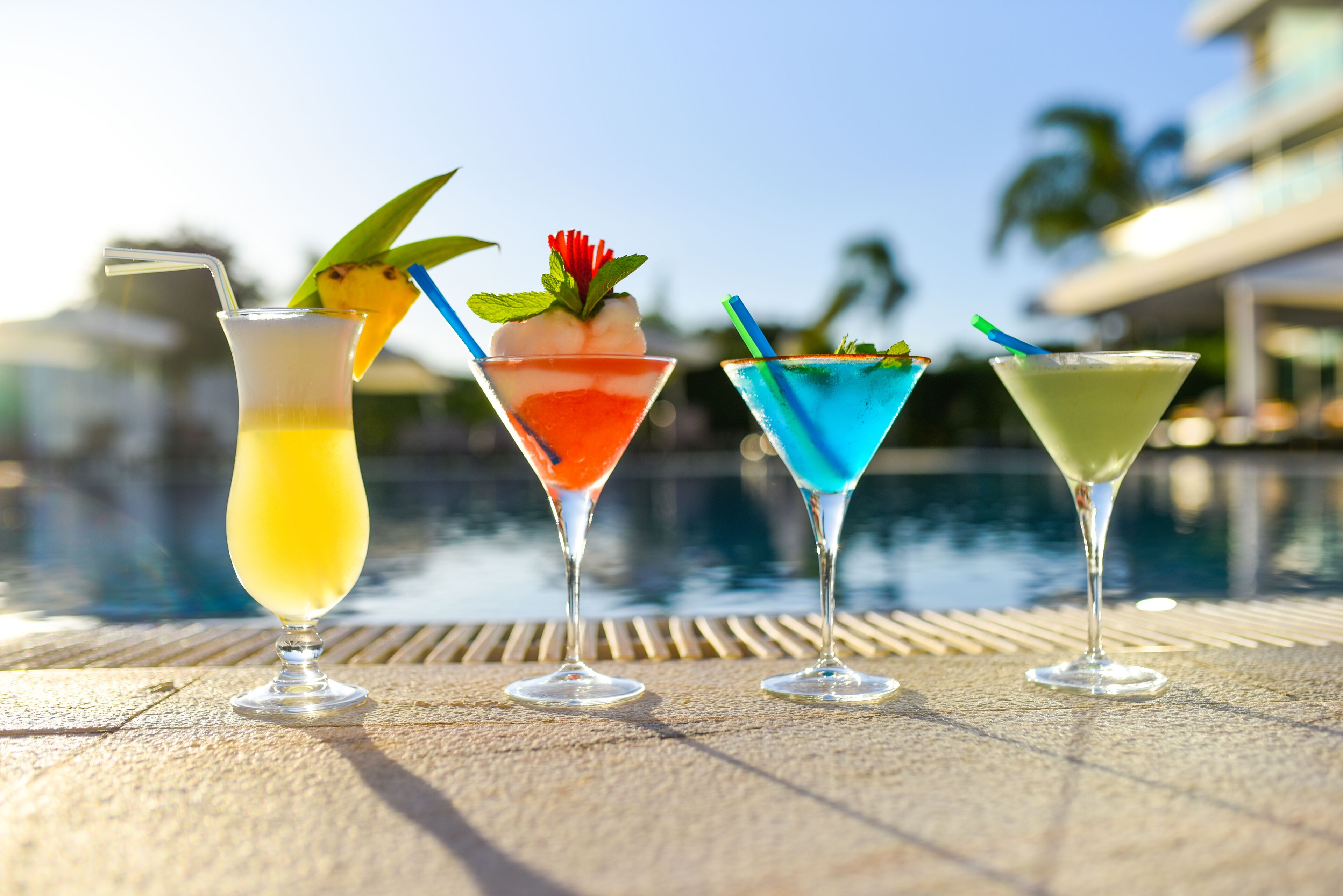 Quatro cocktails sobre uma mesa junto a uma piscina, com vista clara para a piscina e palmeiras ao fundo.