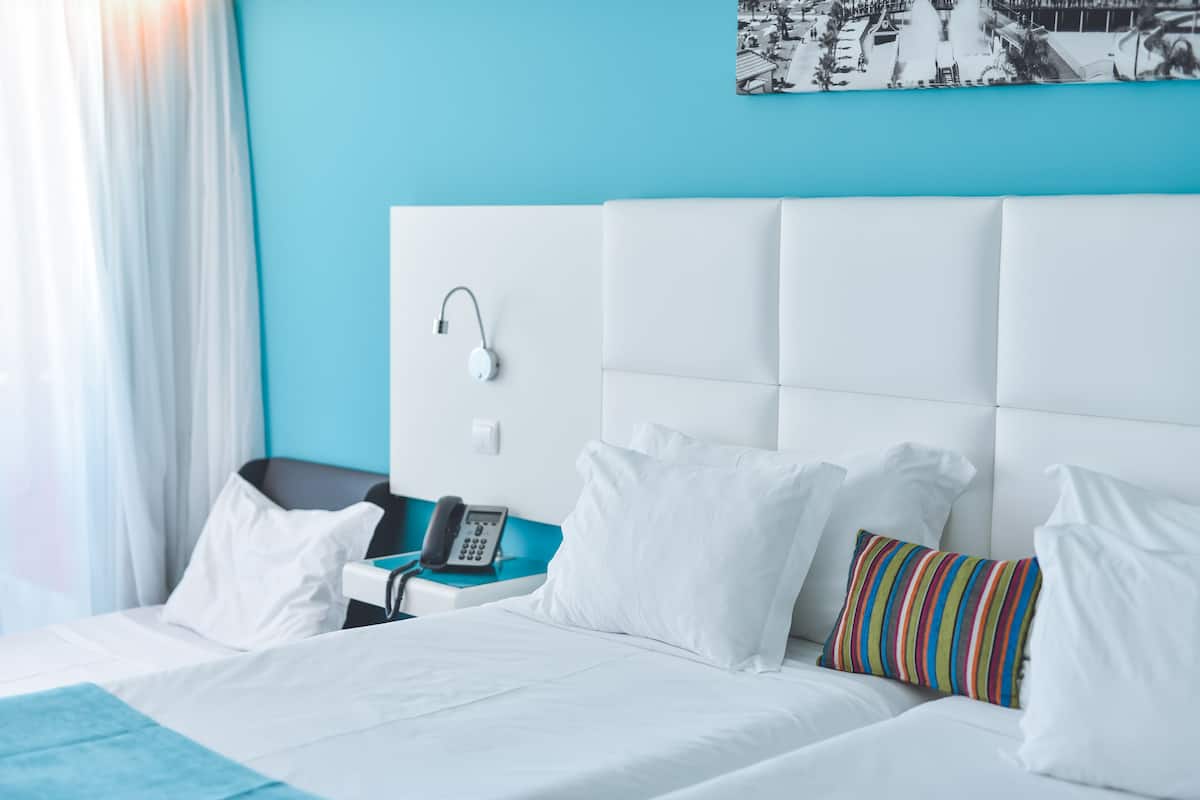 Een hotelkamer met een bed, witte kussens, een kleurrijk gestreept kussen, een wit hoofdbord, een nachtkastje met een telefoon en een blauwe accentmuur.