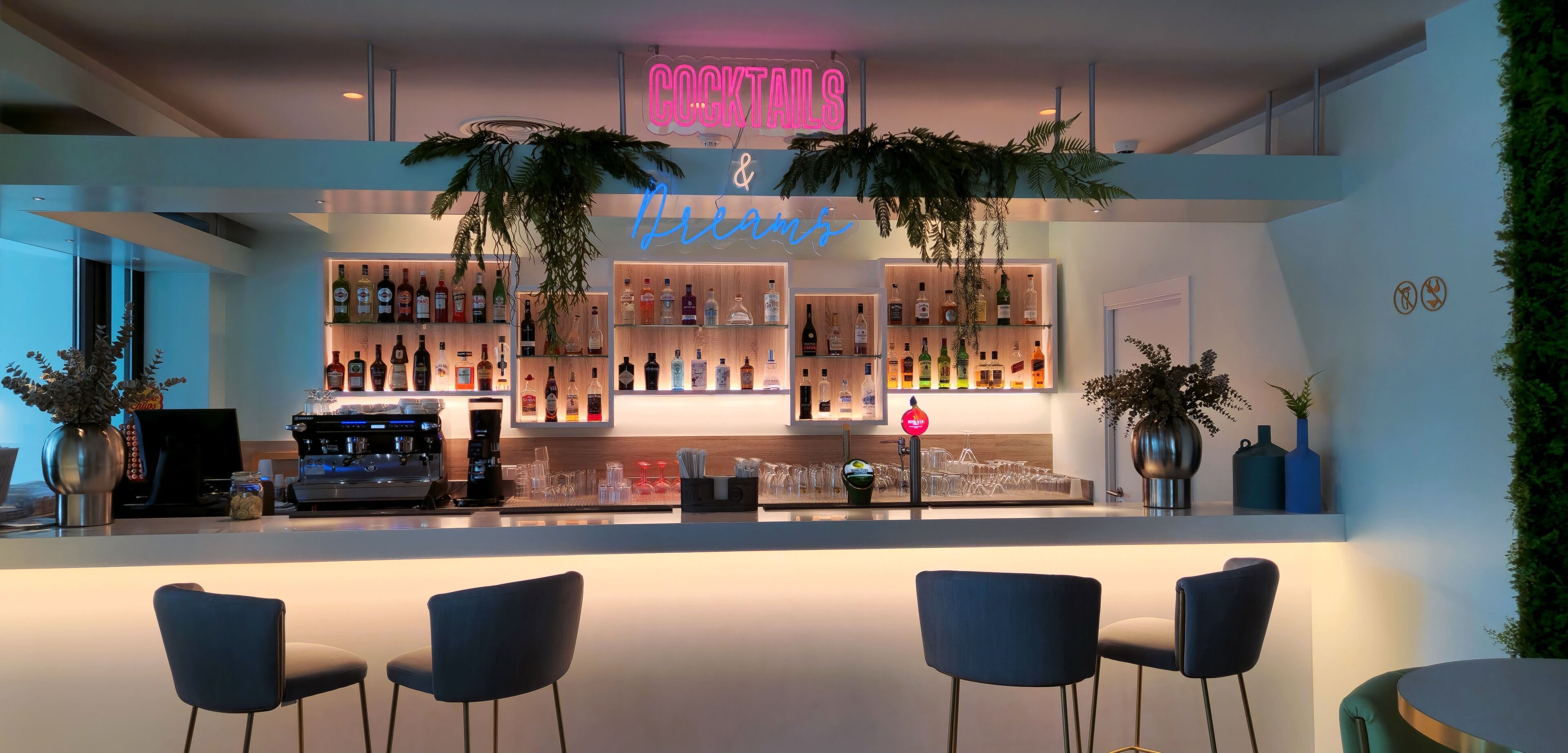 A bar area with a neon sign reading "COCKTAILS & Dreams," shelves of liquor bottles, a coffee machine, and two potted plants.