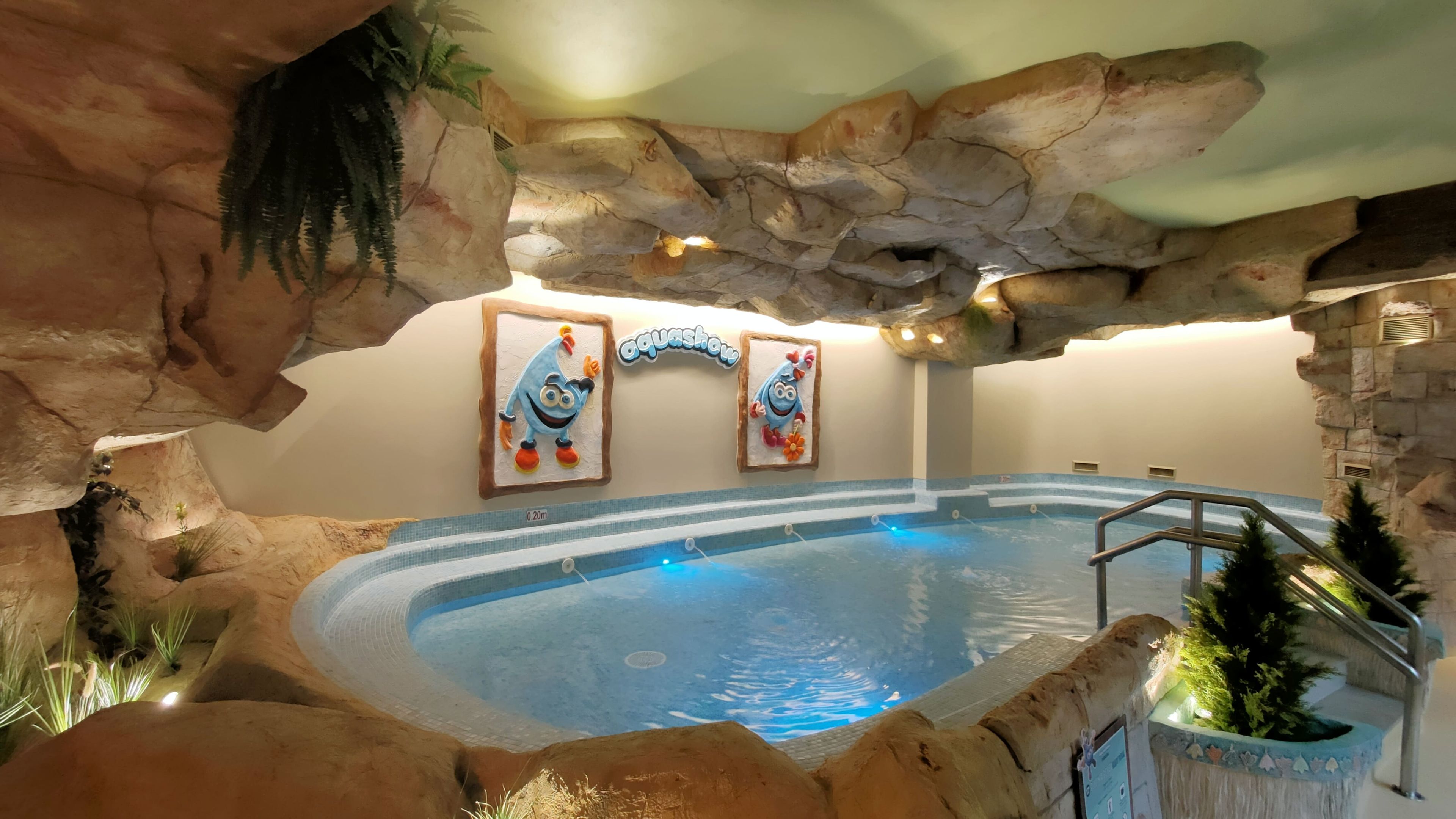 Una piscina interior con una estructura tipo cueva rocosa, un jacuzzi y dos personajes de caricatura enmarcados en la pared.