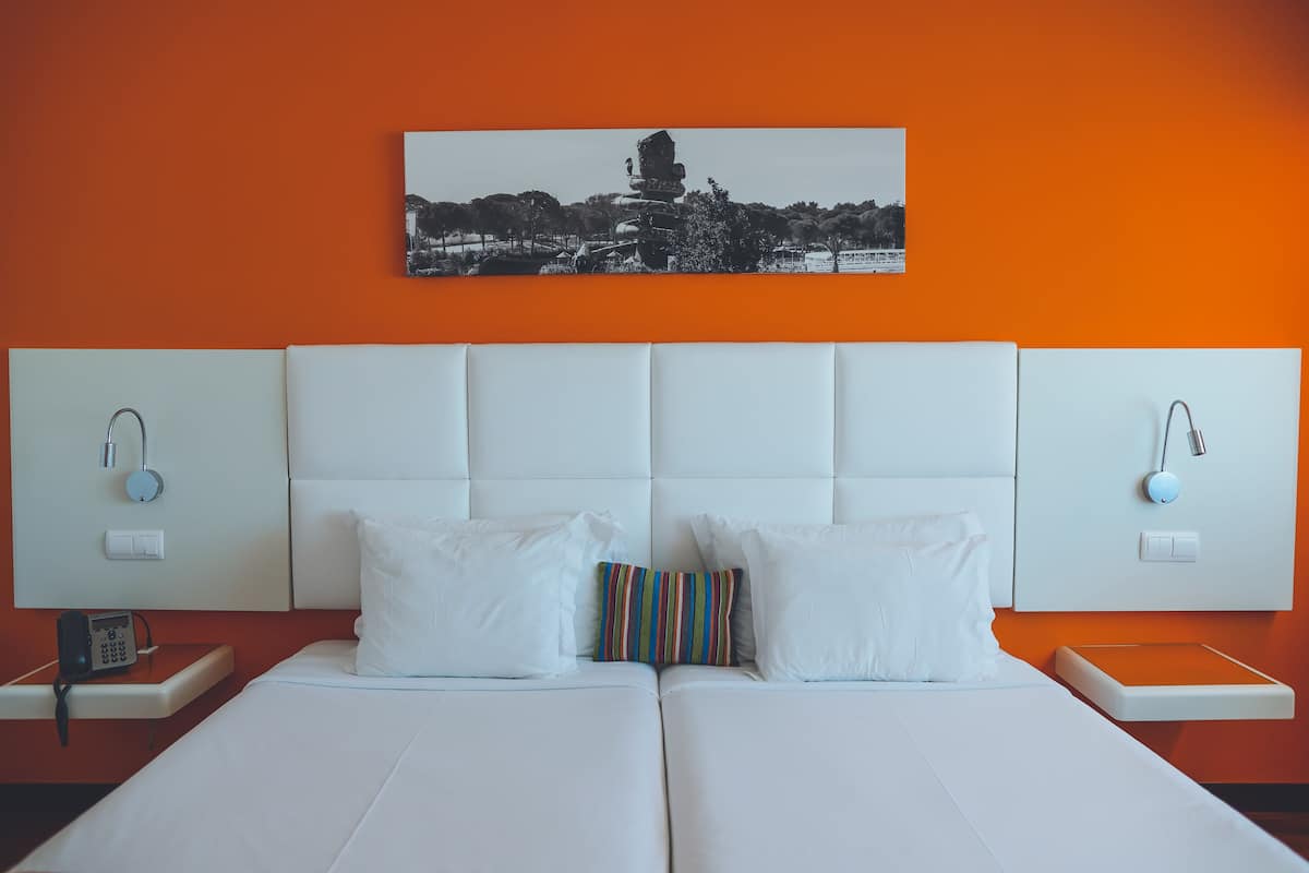 Een tweepersoonsbed met witte beddengoed, twee witte kussens en een kleurrijk gestreept kussen, met een aan de muur hangend beeld van een gebouw.