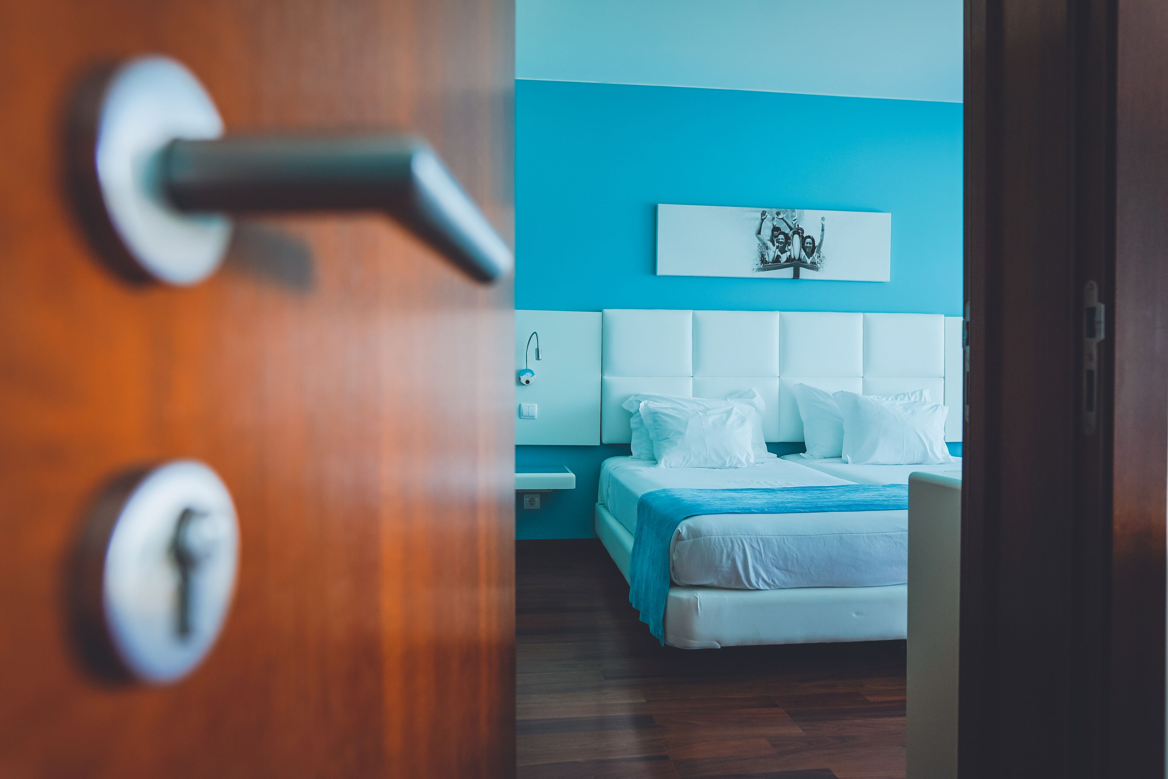 A hotel room with a wooden door, a white bed with a blue blanket, a white headboard, and a wall-mounted artwork.
