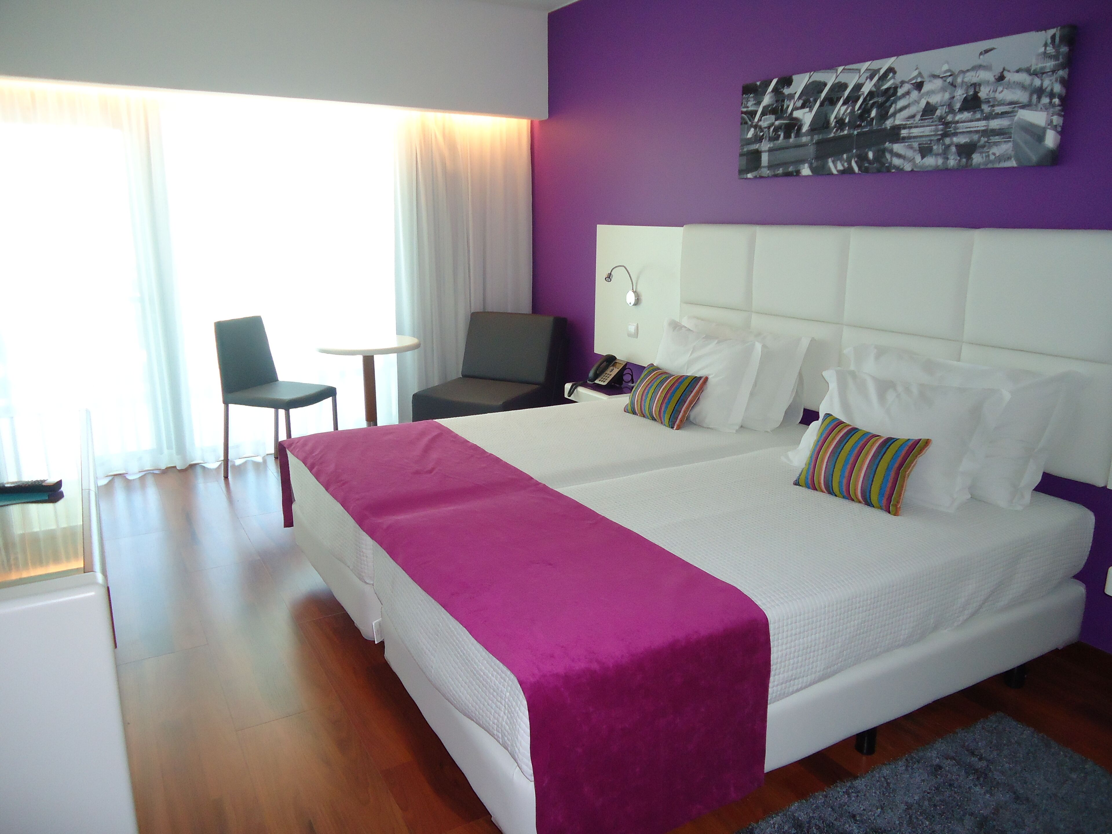 A modern hotel room with a large bed, a small desk, a chair, and a window with curtains.