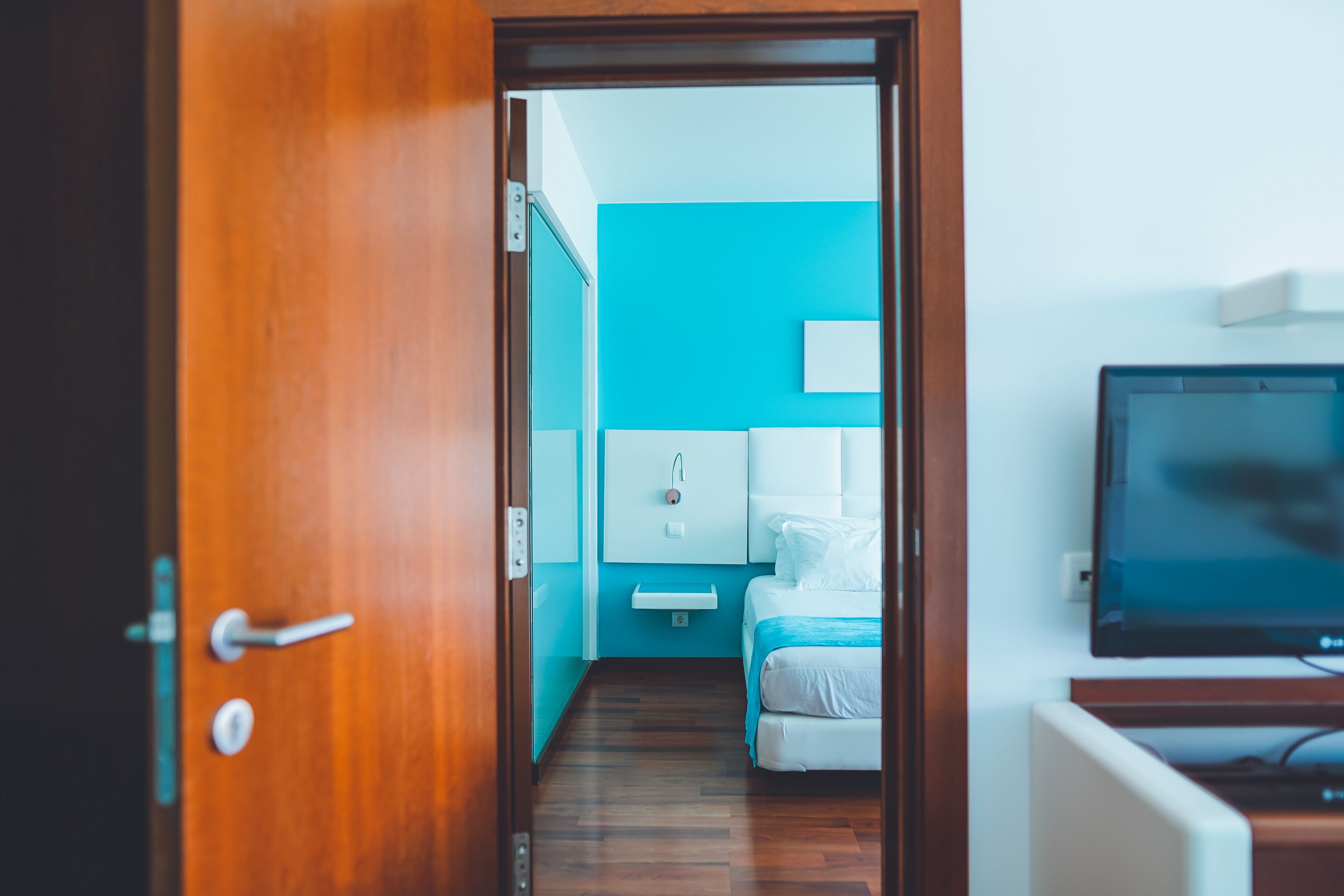 Habitación de hotel con puerta de madera, cama con ropa blanca, mesita de noche, televisor y pared azul.