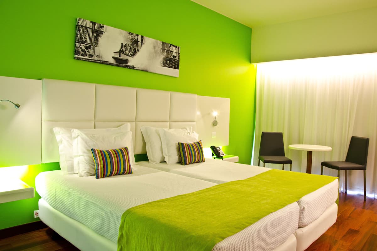 Een hotelkamer met een groene muur, een bed met witte lakens, twee stoelen, een klein tafeltje en een schilderij boven het bed.