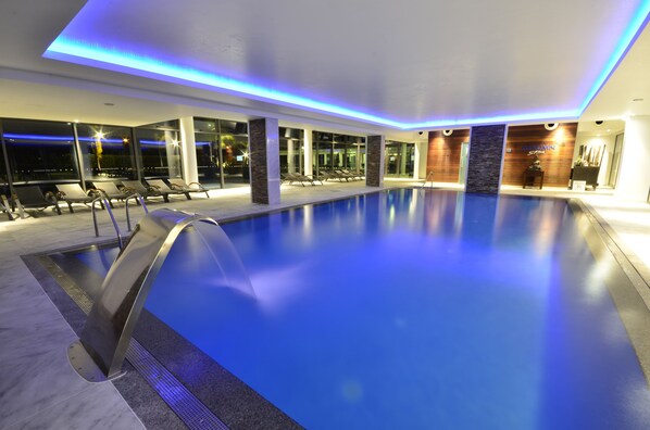 Una piscina interior de diseño moderno, con superficie de agua cristalina, zona de estar junto a la piscina con sillones de descanso e iluminación ambiental azul en el techo.