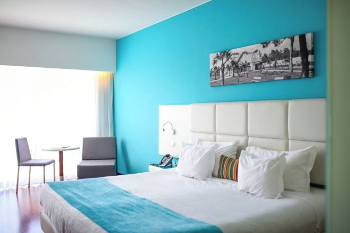 A hotel room with a large bed, a small table, a chair, and a painting on the wall.