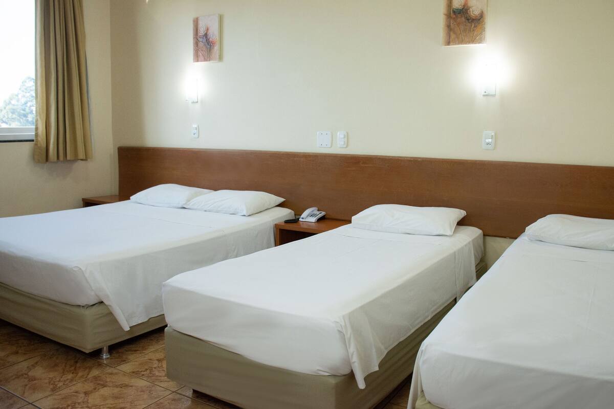 A hotel room with three single beds, a wooden headboard, a bedside table with a phone, and a window with curtains.