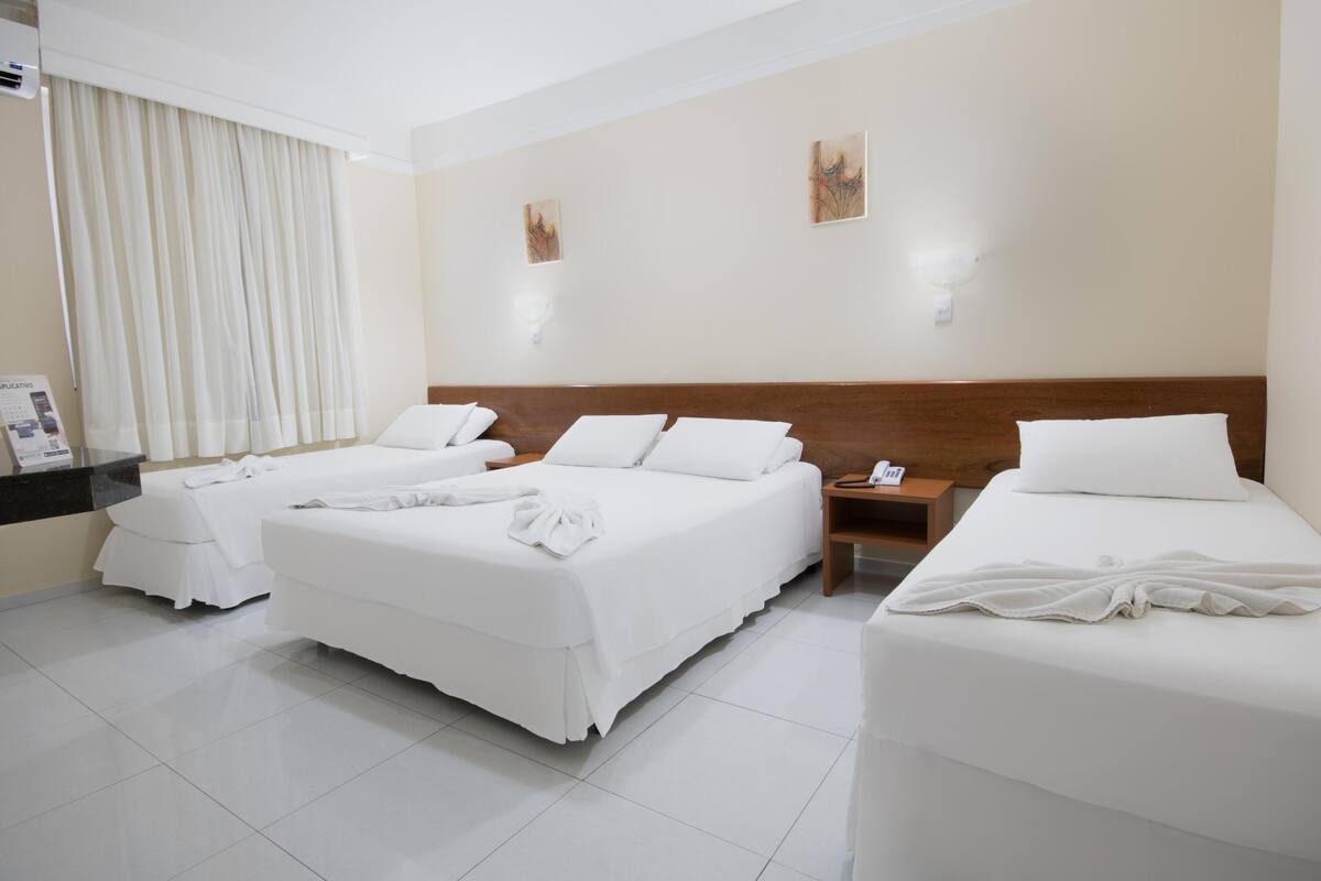 A hotel room with two beds, a bedside table, a nightstand, a window with curtains, and artwork on the wall.