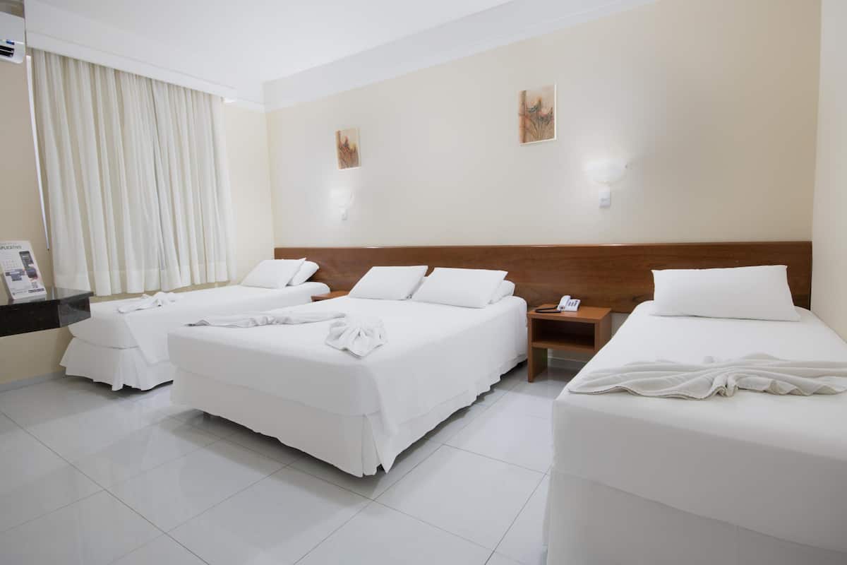 A hotel room with two beds, a bedside table, a nightstand, a window with curtains, and artwork on the wall.
