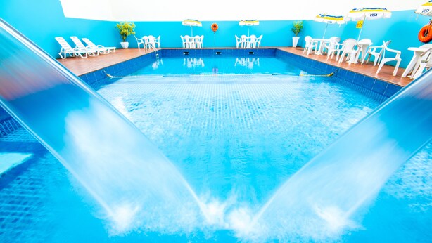 An indoor swimming pool with clear blue water, white lounge chairs, and tables with umbrellas. - Hotel Dan Inn Express Foz do Iguaçu By Nacional Inn (Foz do Iguaçu)