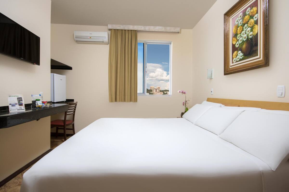 A hotel room with a bed, a desk with a chair, an air conditioner, a window with curtains, and a painting on the wall.