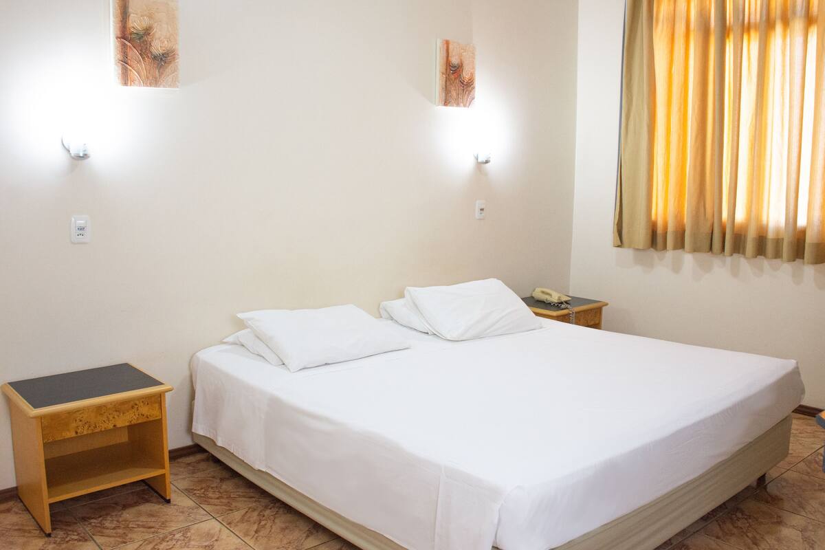 A neatly made bed with white linens, a bedside table, a nightstand, a telephone, and a window with curtains.