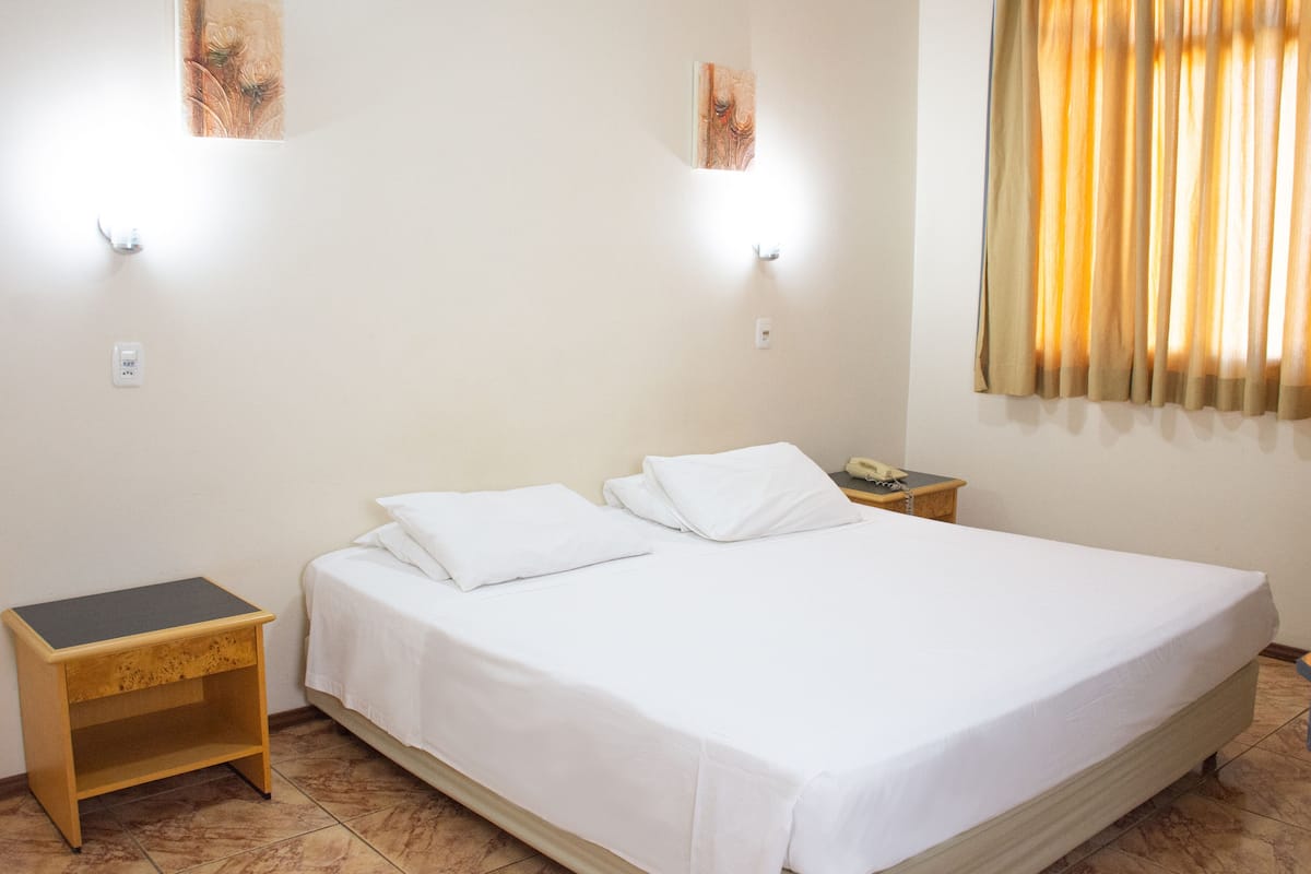 A neatly made bed with white linens, a bedside table, a nightstand, a telephone, and a window with curtains.