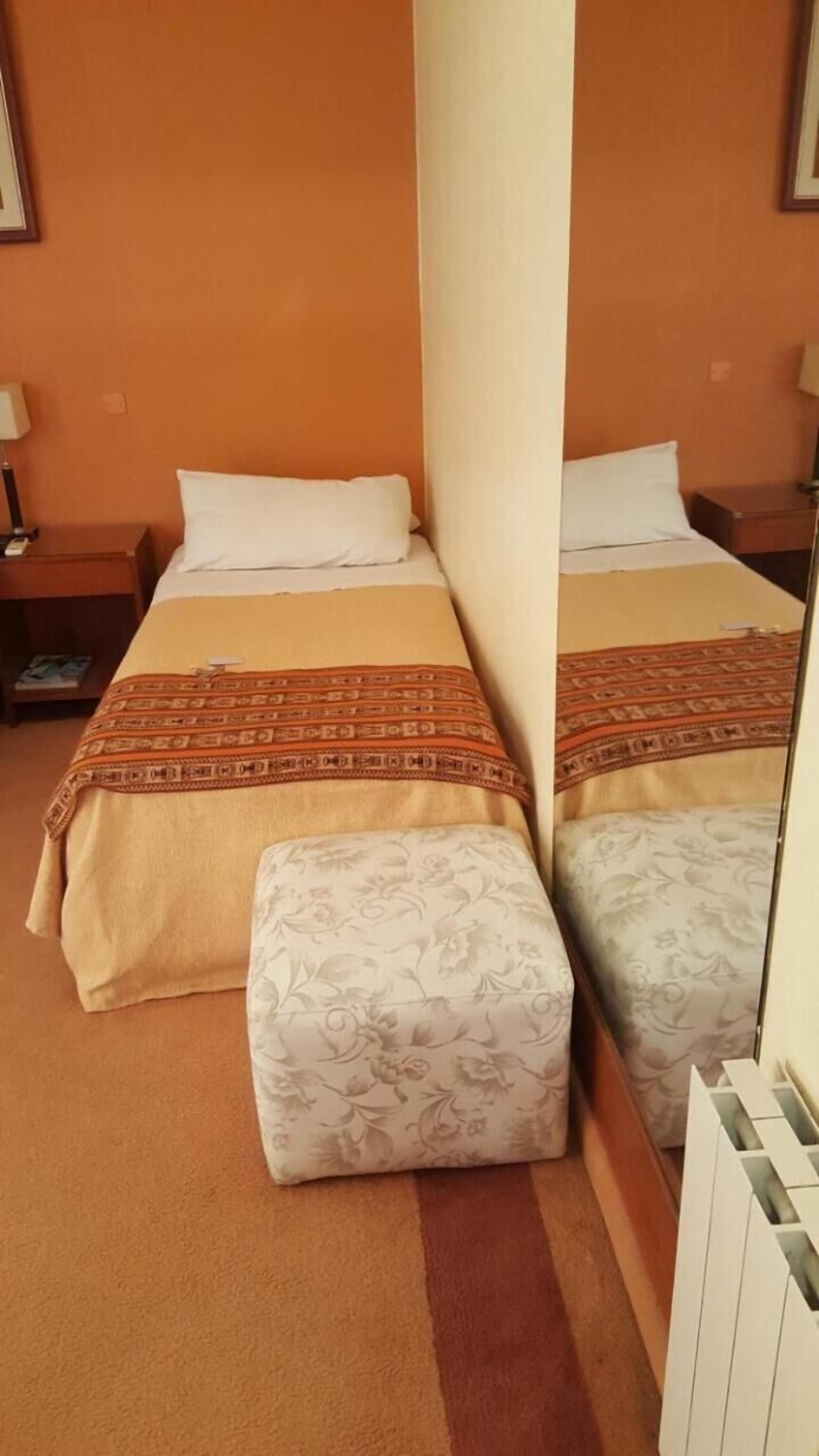 Ein Hotelzimmer mit zwei Betten, jeweils mit gemustertem Bettbezug und Kissen. Ein kleiner Nachttisch mit Lampe und Buch steht daneben. Die Wände sind orange und der Boden ist mit Teppich ausgelegt.