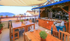 An outdoor dining area with wooden tables and chairs, a buffet with various food items, and a view of the sea.