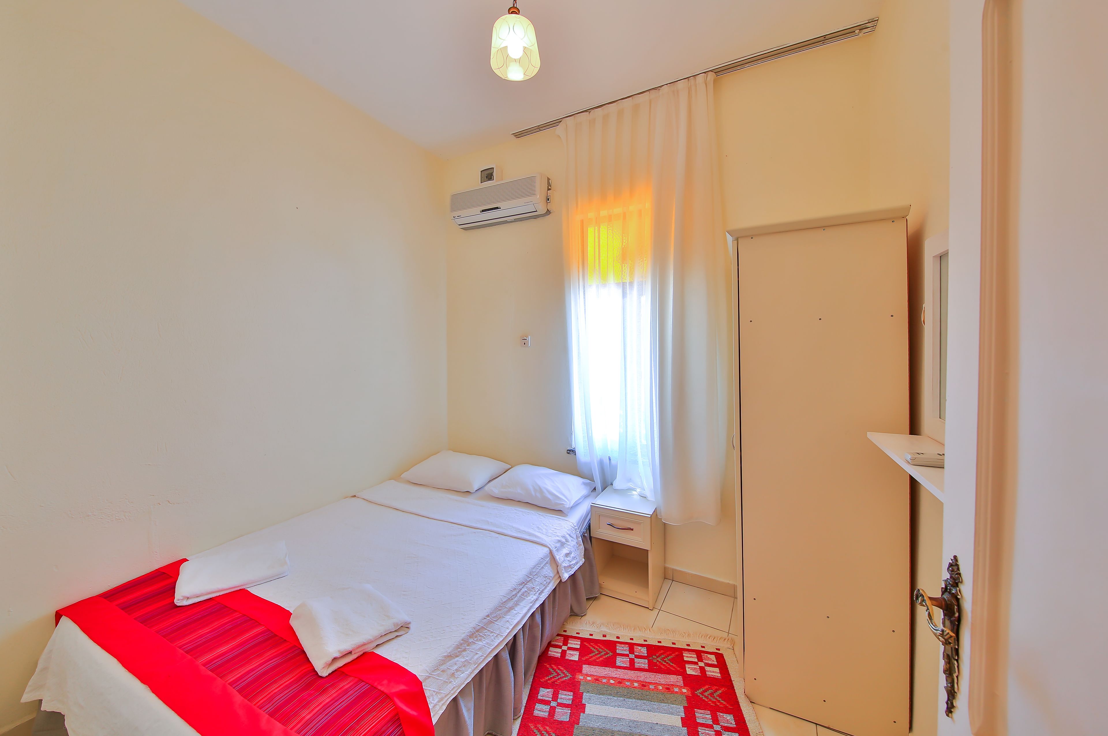 Una habitación pequeña y limpia con una cama individual, una mesita de noche, una ventana con cortinas, un aire acondicionado y una puerta.