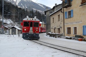 A red train on tracks in a snowy village with buildings and mountains in the background. - Hotel delle Alpi (Sondalo)