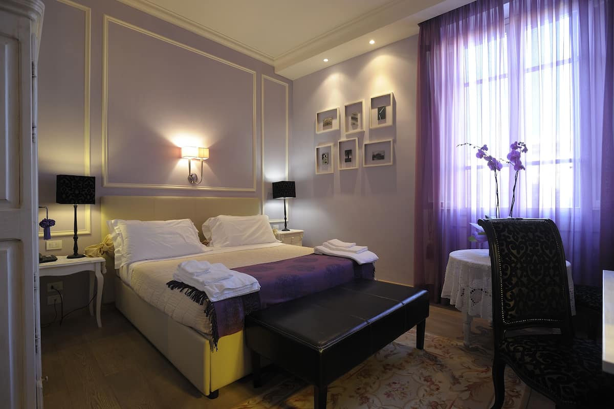 Een hotelkamer met een bed, nachtkastjes, een bankje, een stoel en een raam met gordijnen.