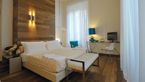 A modern hotel room with a wooden accent wall, a large bed with a quilted duvet, a desk with a computer, and a blue chair.