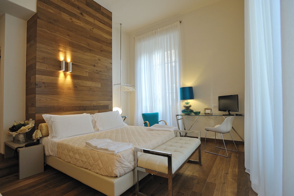 Een moderne hotelkamer met een houten accentmuur, een groot bed met een gewatteerde spreideken, een bureau met een computer en een blauwe stoel.