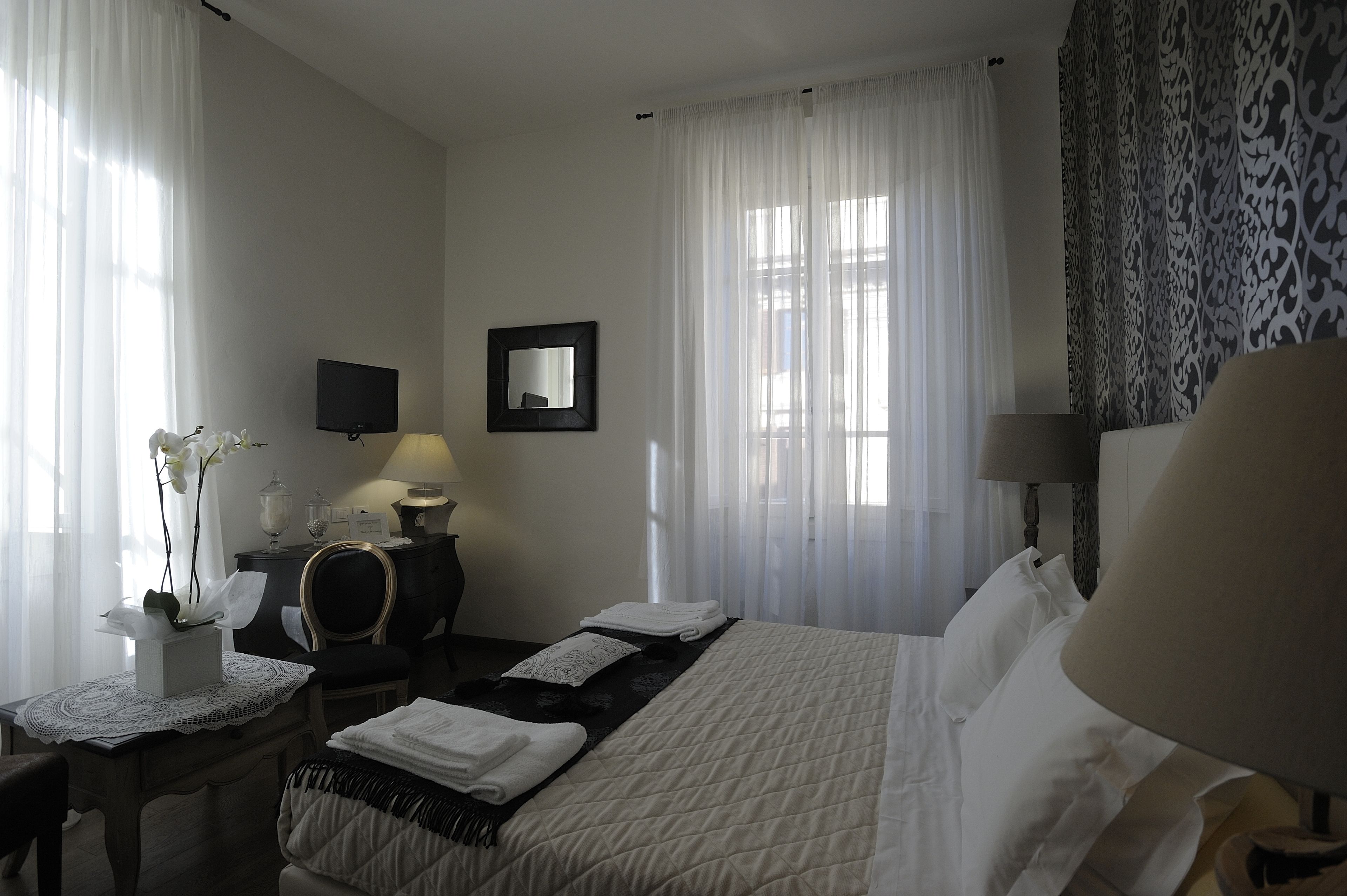A hotel room with a bed, a desk with a lamp, a chair, a TV, and a window with sheer curtains.