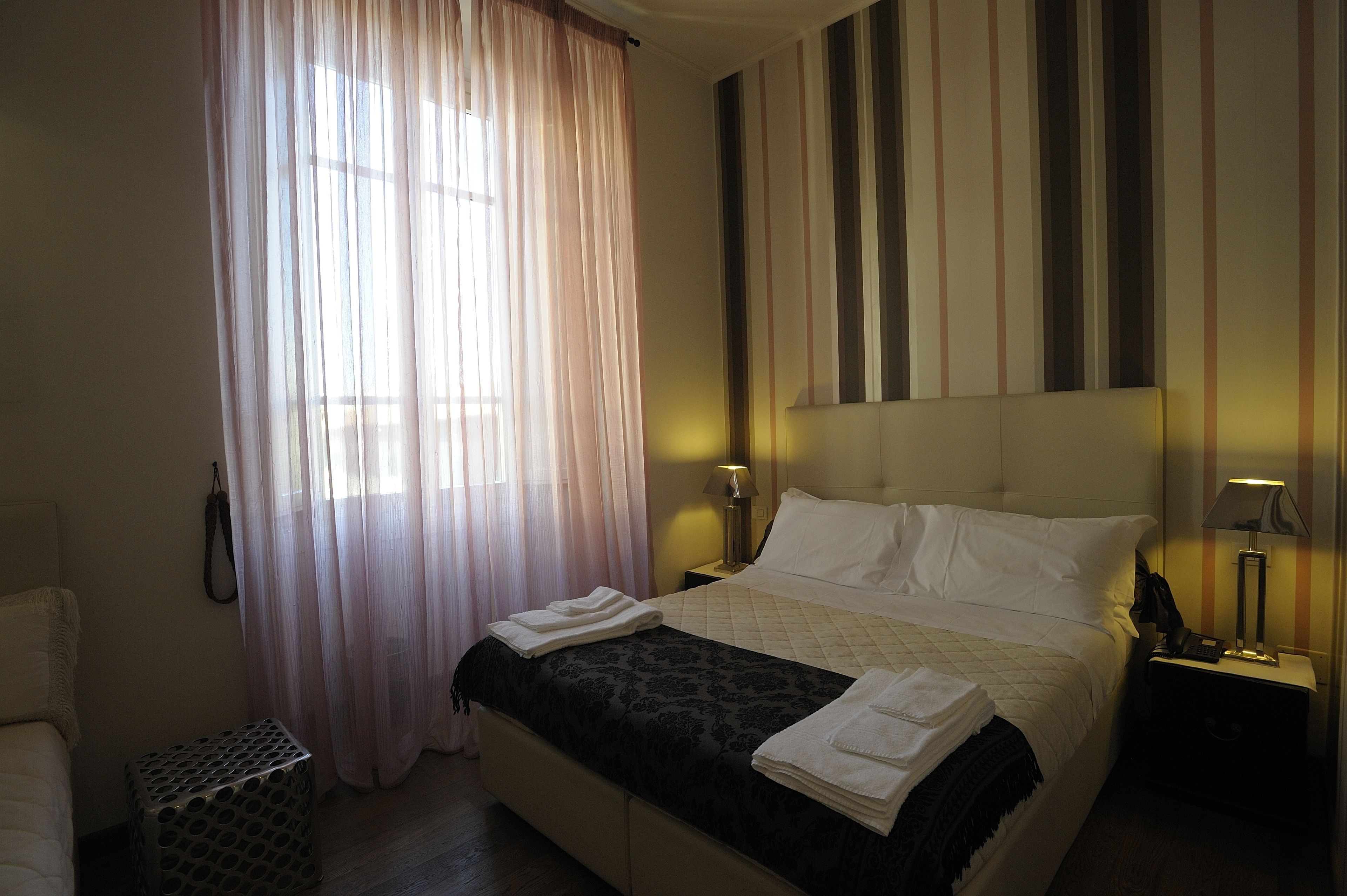 A hotel room with a bed, a bedside table with a lamp, a small table with a patterned cover, and a window with sheer curtains.