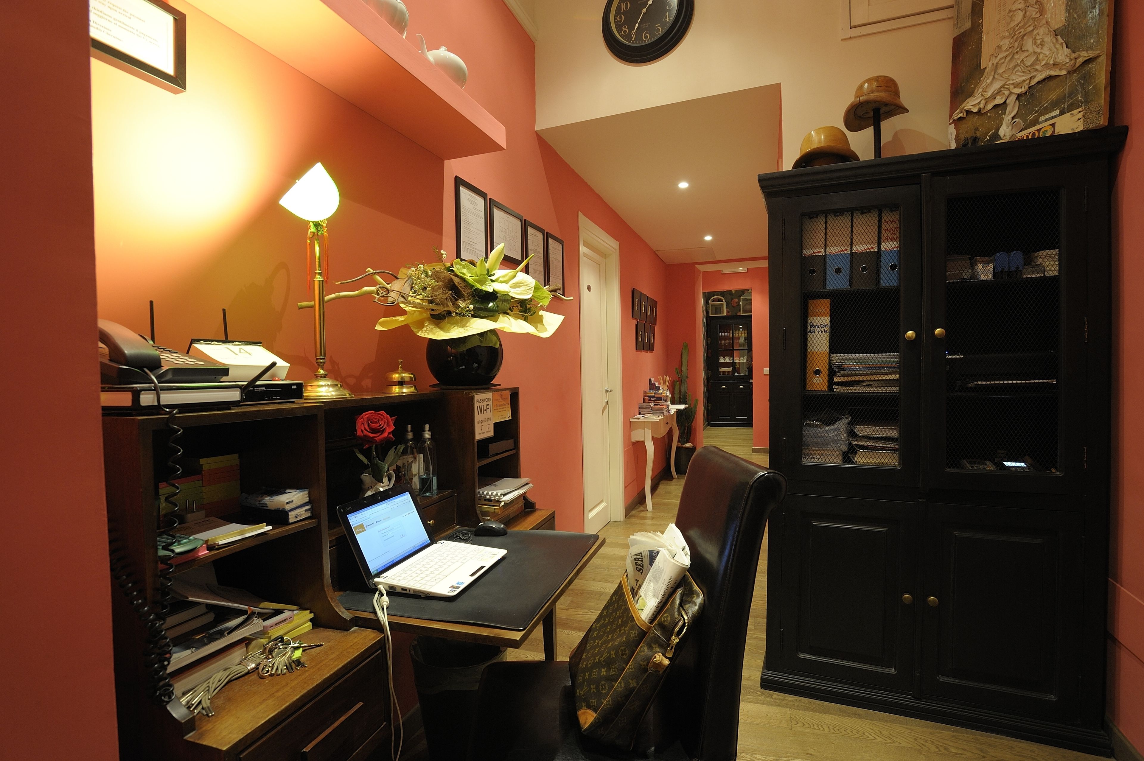 Ein Hotelzimmer mit Schreibtisch, Laptop, Lampe und einer Zimmerpflanze. Es gibt einen schwarzen Schrank mit Glastüren und eine Uhr an der Wand.