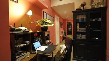 Ein Hotelzimmer mit Schreibtisch, Laptop, Lampe und einer Zimmerpflanze. Es gibt einen schwarzen Schrank mit Glastüren und eine Uhr an der Wand.